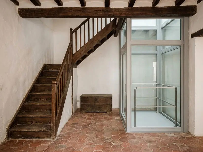 Elevador de casa con cabina abierta y barandilla junto a una escalera de madera